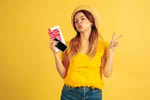 Free photo tickets, gadget. caucasian woman's portrait on yellow studio background. beautiful female model in hat. concept of human emotions, facial expression, sales, ad. summertime, travel, resort concept.