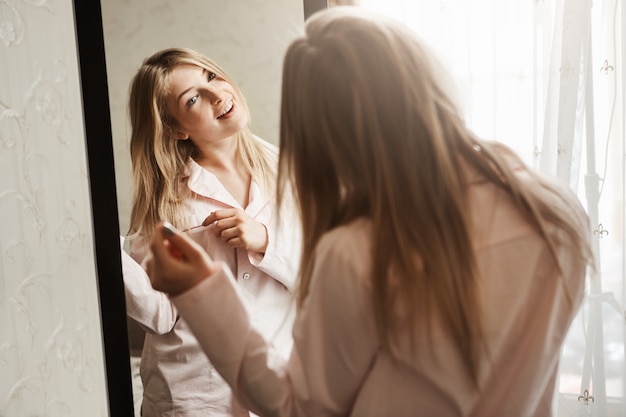 Free Photo time to dress up and went to meet adventures. home shot of beautiful blond caucasian girl looking in mirror, wearing nightwear and touching hair strand, thinking about new hairstyle