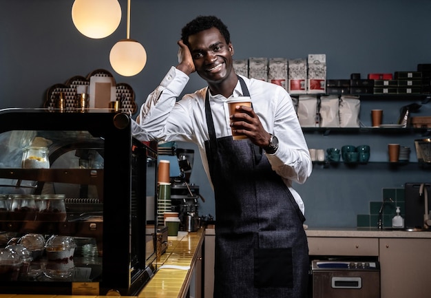 Free Photo tired african barista holding a cup with coffee while leaning on a counter in a coffee shop and looking at a camera with a happy look .