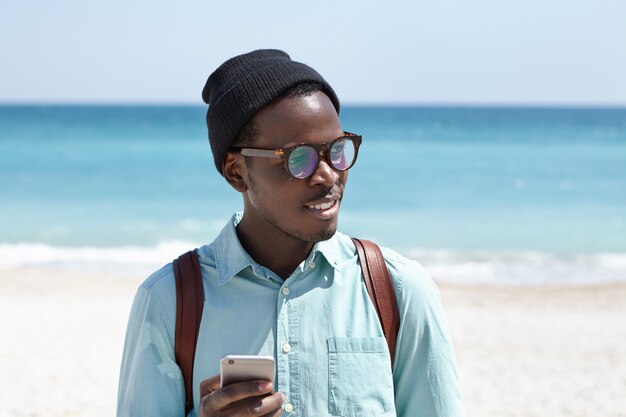 Tired Afro American backpacker in hat and eyewear using online taxi service app on mobile phone to request cab while feeling thirsty, looking for place to have some cold drink