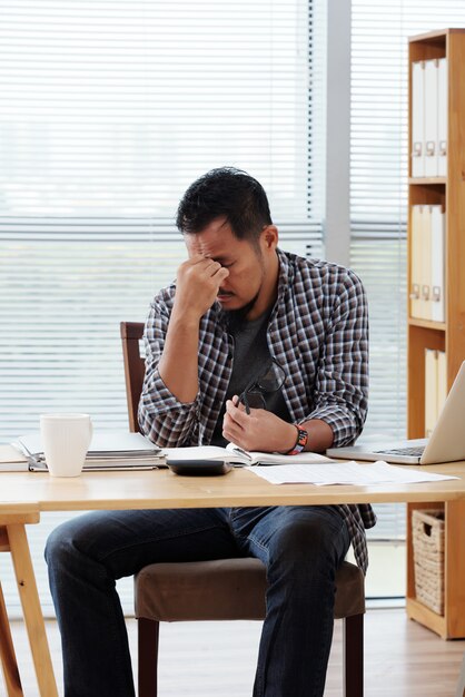 Tired Asian entrepreneur sitting at table in office and rubbing his forehead