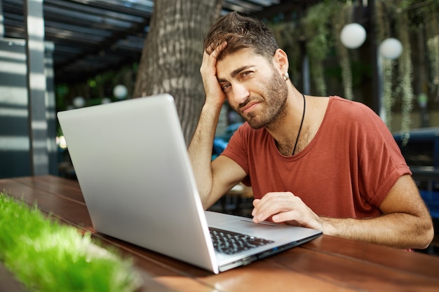 Tired bearded handsome freelancer, male programmer or student using laptop while sitting outdoor cafe