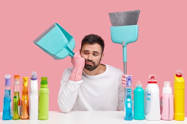 Tired bearded man carries broom, scoop, feels tired after sweeping and cleaning floor, sits at workplace with detergents