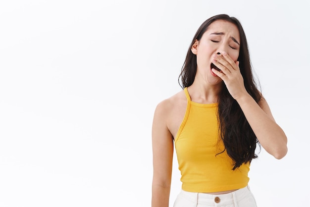 Tired or bored attractive stylish brunette eastasian woman exhausted of uninteresting conversation falling asleep from boredom yawning tilt head and cover opened mouth stand white background