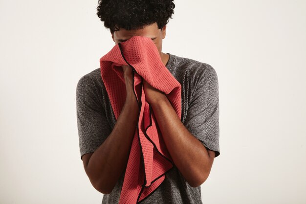 Tired disappointed young fit black athlete in gray shirt wiping sweat from his face with red waffle towel on white