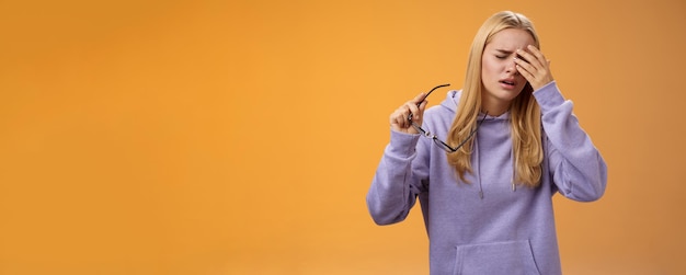 Tired dizzy cute blond girl taking off glasses touching forehead feel sick unwell suffering headache painful migraine standing bothered uncomfortable orange background in hoodie