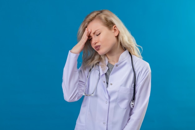 Tired doctor young girl wearing stethoscope in medical gown put hand on forehead on blue background