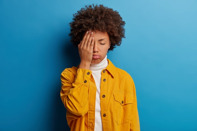 Tired fed up woman covers half of face, keeps eyes closed, sighs from tiredness, feels exhausted to work without rest, poses in studio against blue wall