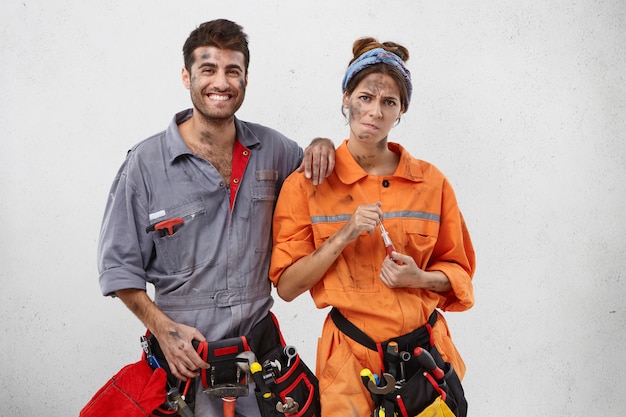 Free photo tired female carpenter in orange uniform, holds screwdriver and male colleague with happy expression