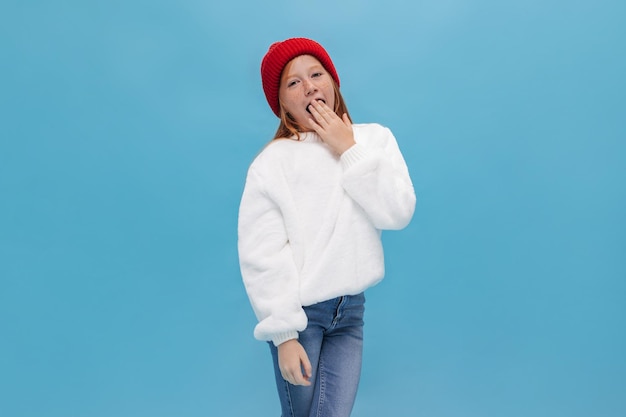 Free Photo tired ginger girl with freckles in oversize shirt and jeans yawns on blue background young cute lady in red hat posing on isolated backdrop