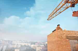 Free photo tired and handsome builder in hat lying on brick wall on high and resting. looking away. blue sky with clouds at summer season on background. milk and bread near.