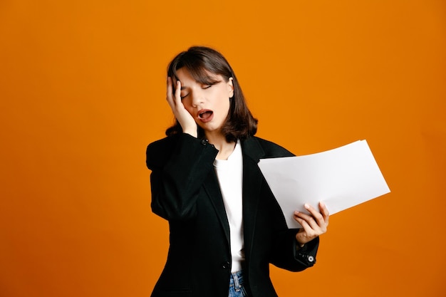 tired holding papers young beautiful female wearing black jacket isolated on orange background