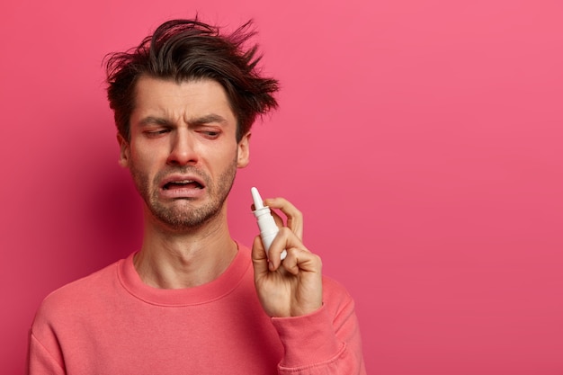 Free photo tired ill man has symptomes of cold, holds nasal spray, wants to recover quickly, uses effective medicine, drips nose, getting worse, isolated on pink wall, feels bad. treatment of flu