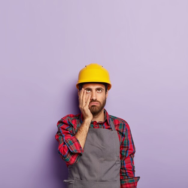 Tired industrial worker wears yellow hardhat and apron