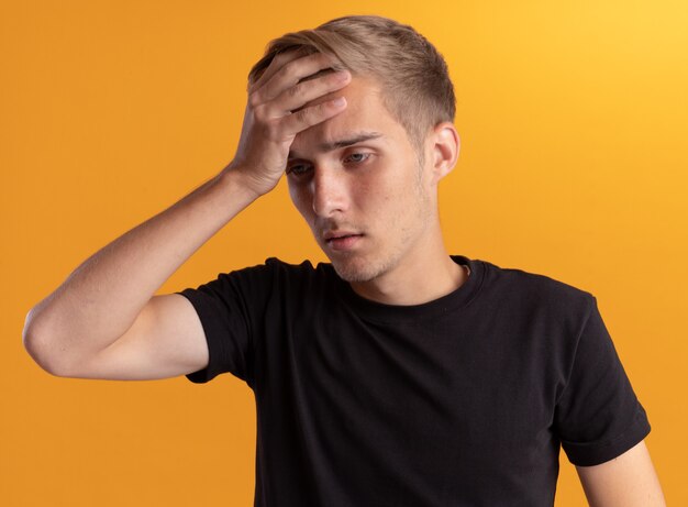 Tired looking at side young handsome guy wearing black shirt putting hand on forehead isolated on yellow wall