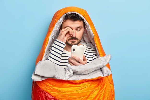 Tired male camper rubs eyes, uses mobile phone, tries to connect to intenrnet in wild nature, poses in sleeping bag, has all necessary equipment needed for camping