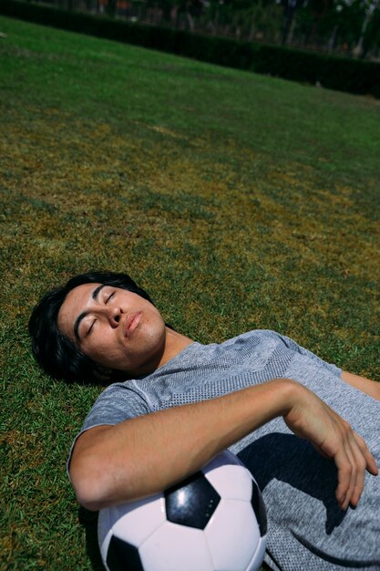 Tired man lying with eyes closed on grass with football