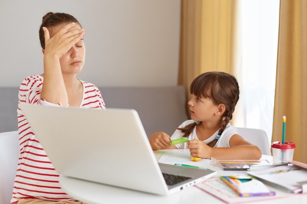 Tired nervous female wearing striped casual shirt being tired to explains home task, being exhausted, covering eyes with palm, child looking at mother, online education.