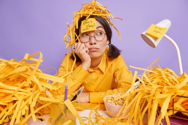 Free photo tired onworked student prepares for examination session sits at desktop with thoughtful expression surrounded by piles of cut paper makes notes on stickers not to forget about necessary tasks