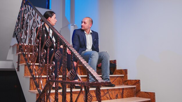 Tired overworked businesspeople taking a break sitting on stairs in corporate business office discussing. Colleague entrepreneur doing overtime together late at night making plans for deadline project