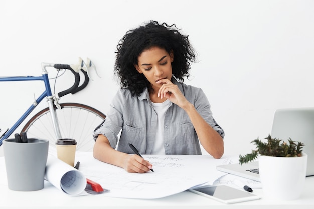 Tired serious young dark-skinned woman engineer looking at her drawing