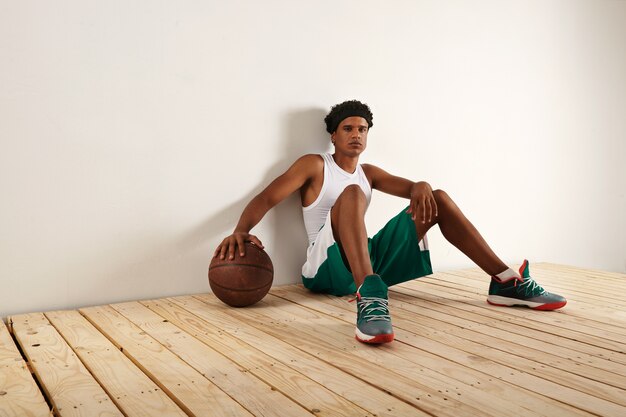 Tired and thoughtful black basketball player in green and white basketball outift sitting on light wooden floor resting his hand on a grunge brown basketball