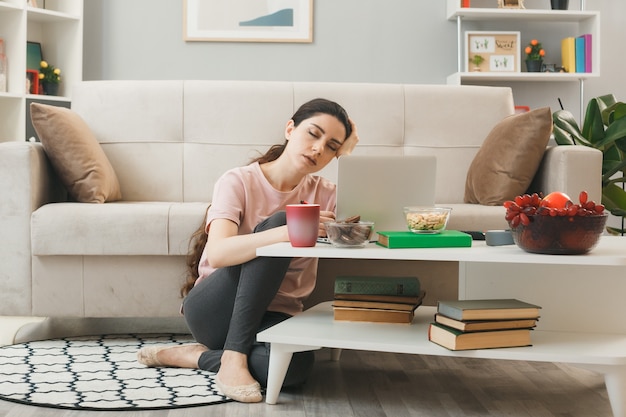 Tired with closed eyes putting hand on head young girl sitting on floor behind coffee table in living room