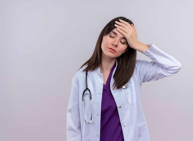 Tired young female doctor in medical robe with stethoscope puts hand on head on isolated white background with copy space