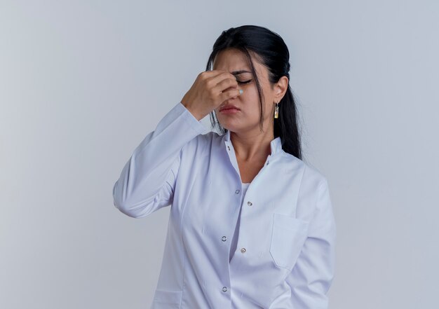 Tired young female doctor wearing medical robe holding nose with closed eyes isolated on white wall with copy space