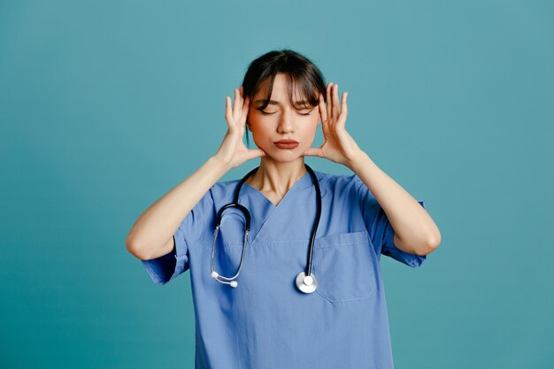 Tired young female doctor wearing uniform fith stethoscope isolated on blue background