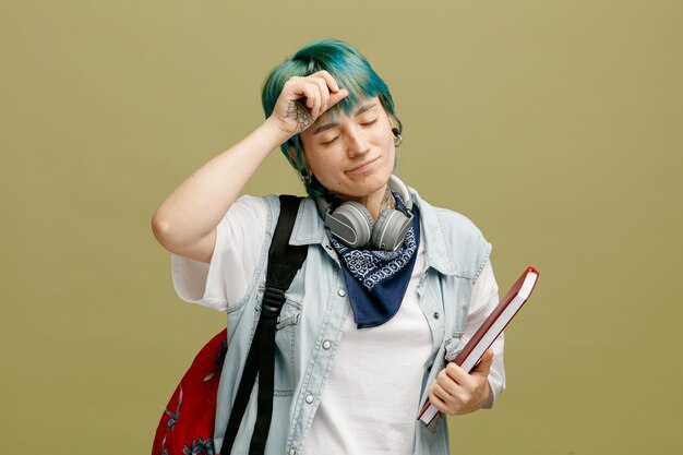 Free photo tired young female student wearing headphones and bandana on neck and backpack holding note book touching head with closed eyes isolated on olive green background