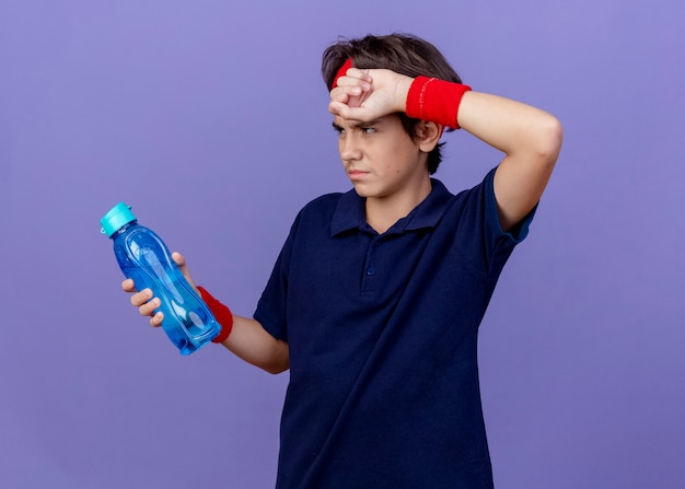 Tired young handsome sporty boy wearing headband and wristbands with dental braces putting hand on forehead holding and looking at water bottle isolated on purple wall