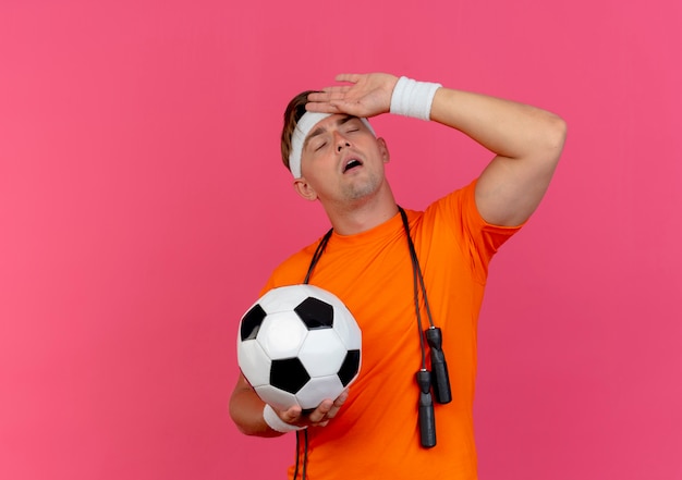 Free photo tired young handsome sporty man wearing headband and wristbands with jump rope around neck holding soccer ball putting hand on forehead with closed eyes isolated on pink background with copy space