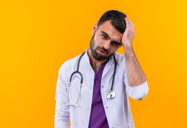 Tired young male doctor wearing stethoscope medical gown put his hand on head on isolated yellow wall