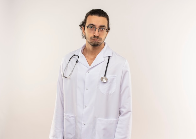 Tired young male doctor with optical glasses wearing white robe with stethoscope on isolated white wall with copy space