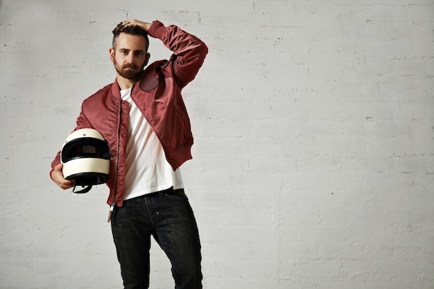 Tired young motorbiker in red bomber jacket, black jeans and white t-shirt adjusting his hairstyle and holding his white helmet