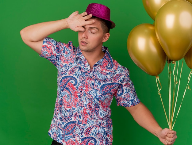 Free photo tired young party guy with closed eyes wearing pink hat holding balloons and putting wrist on forehead isolated on green