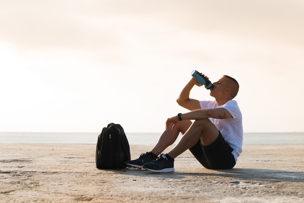 Tired young sportsman drinking water on ground