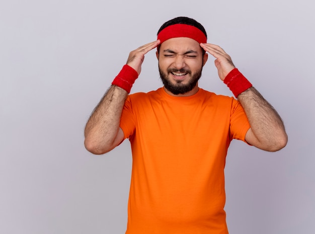 Free photo tired young sporty man with closed eyes wearing headband and wristband putting fingers on temple isolated on white background with copy space