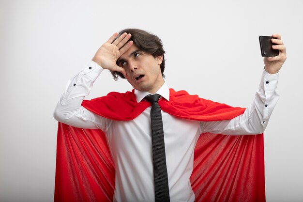 Tired young superhero guy wearing tie take a selfie and putting hand on head isolated on white