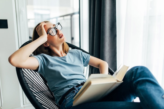 Free photo tired young woman with book sleeping on lounge chair at home