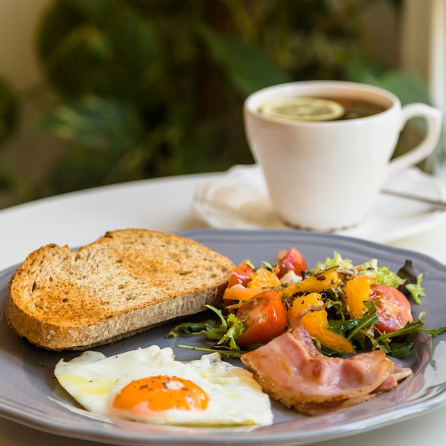 Toast; half fried egg; salad and bacon on gray plate near the tea cup