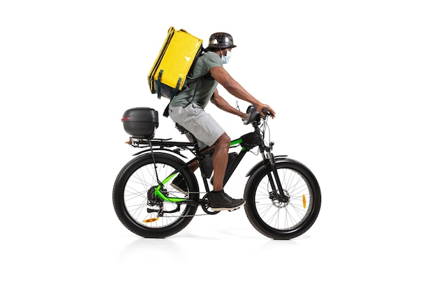 Too much orders. Contacless delivery service during quarantine. Man delivers food during isolation, wearing helmet and face mask. Taking food on bike isolated on white wall. Safety. Hurrying up.