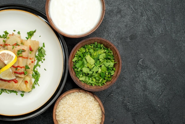 Free Photo top close-up side appetizing dish stuffed cabbage with lemon herbs and sauce on white plate and sour cream rice herbs in bowls on the left side of dark table