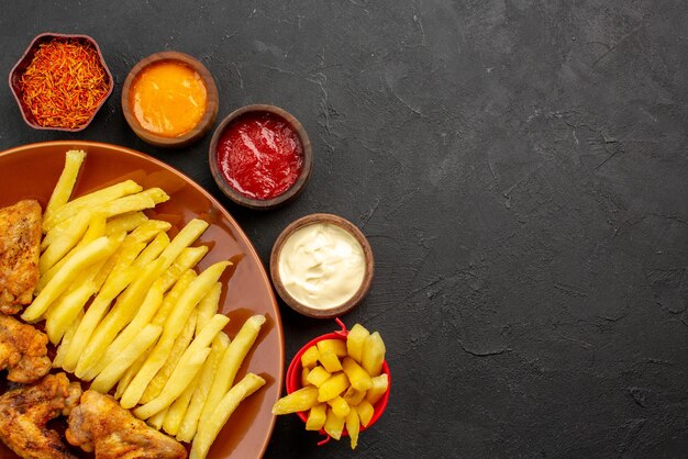 Top close-up view chiken and potatoes chicken wings french fries and lemon three bowls of different types of sauces and spices on the dark table