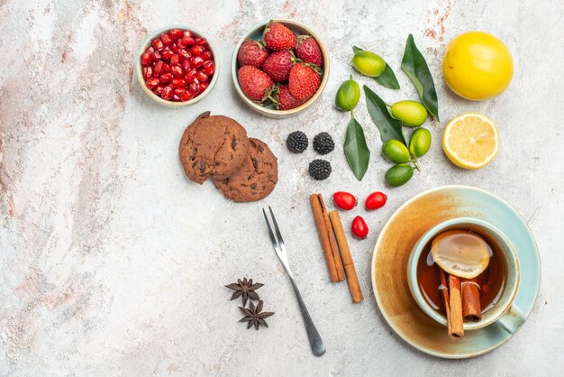 Top close-up view chocolate cookies chocolate cookies a cup of tea with lemon and cinnamon sticks fork bowls of berries citrus fruits pomegranate on the table