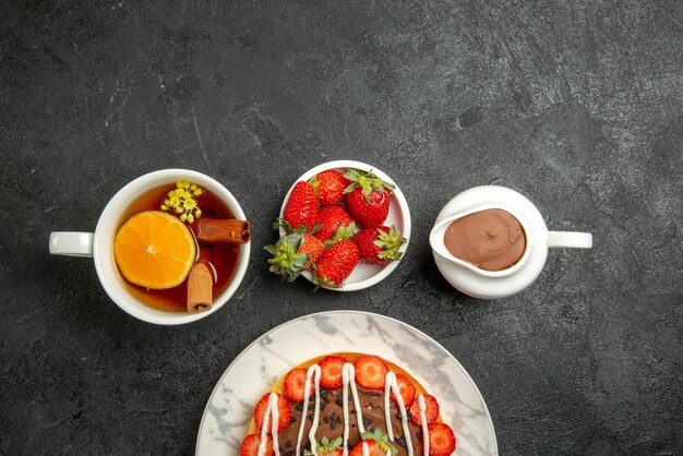 Top close-up view chocolate cream strawberries appetizing cake and a cup of tea with lemon and cinnamon next to the bowls of strawberries and chocolate cream on the dark table