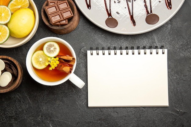 Top close-up view citrus fruits chocolate bowls of citrus fruits and chocolate next to the white notebook and a cup of tea with lemon