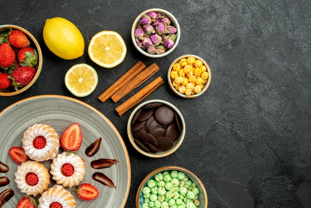 Top close-up view cookies with chocolate and strawberry different sweets with black tea lemons hizelnuts bowls of chocolate on the left side of table