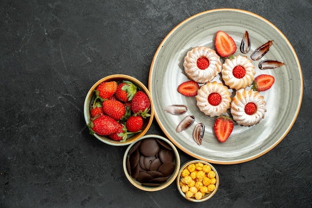 Free photo top close-up view cookies with strawberry bowls of chocolate strawberry and hizelnuts next to appetizing cookies with chocolate and strawberry on black surface